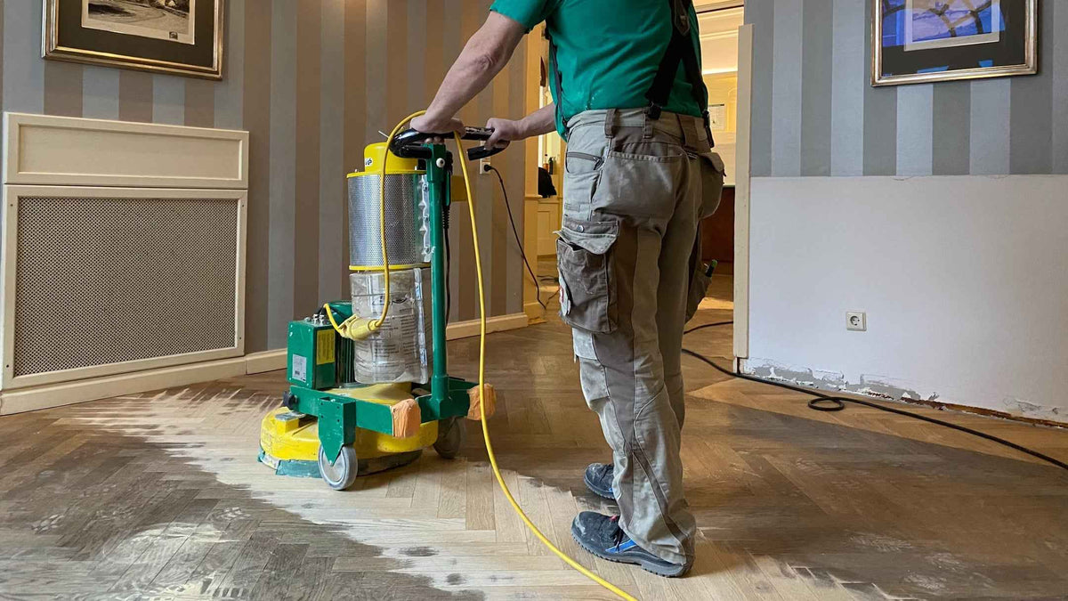 Ein Arbeiter schleift einen Fischgrätparkettboden mit einer großen gelben Schleifmaschine